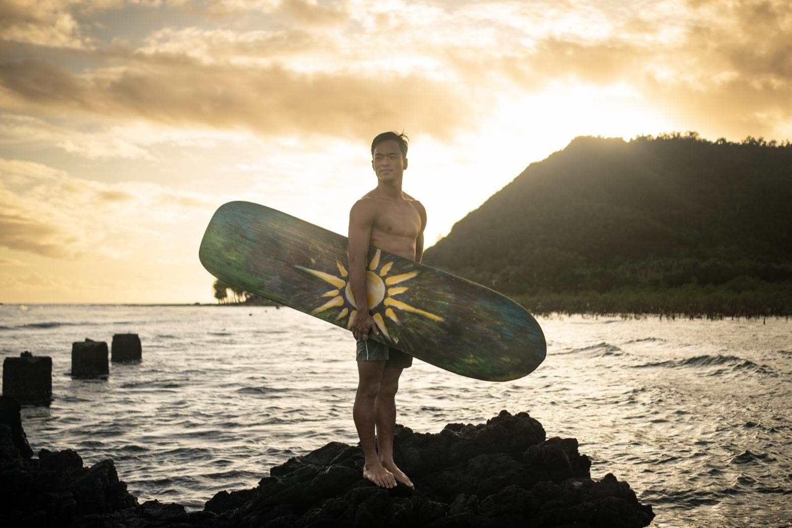 A surfer holding a board on the rocks at sunset